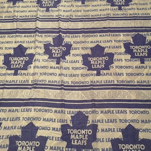 May include: A white and blue patterned fabric featuring the text "TORONTO MAPLE LEAFS" and maple leaf logos. The design is repeated across the fabric, creating a uniform pattern. The fabric is likely cotton or a similar material.