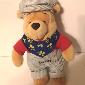May include: A plush Winnie the Pooh bear wearing blue and white striped overalls, a red shirt, a blue bandana with yellow bees, and a blue and white striped hat. The bear is standing on a white surface. The bear's name, "Pooh", is written on the overalls.