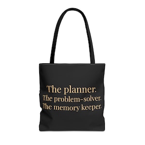 May include: Black tote bag with black handles. The bag features the text "The planner. The problem-solver. The memory keeper." in a gold color. The bag is designed for carrying items.