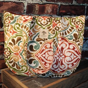 May include: A large tote bag with a colorful floral pattern in shades of red, green, brown, and blue. The bag has two handles and a tag with text. The bag is sitting on a wooden surface in front of a brick wall.
