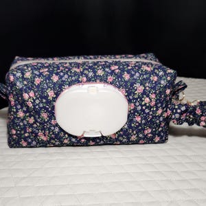 May include: A navy blue floral print wipe case with a white plastic dispenser. The case has a zipper closure and a matching fabric handle. The floral pattern includes pink and white flowers.