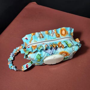 May include: A light blue baby wipes case with a zipper closure and a ruffled edge. The case is decorated with a colorful animal print featuring giraffes, lions, and elephants. It has a matching handle with a metal clasp.