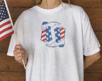 May include: White t-shirt featuring a graphic of two cowboy boots with red, white, and blue American flag designs. The shirt is a relaxed fit and is suitable for casual wear. The person is holding a small American flag.
