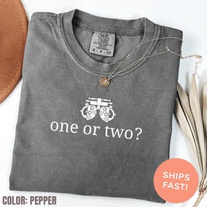 Puede incluir: Una camiseta gris Comfort Colors con el texto "one or two?" y una ilustración de una máquina de examen ocular. Un collar plateado con un colgante está sobre la camiseta. La camiseta está etiquetada como "Color: Pepper".
