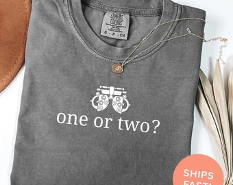 May include: A gray Comfort Colors t-shirt with the text "one or two?" and an illustration of an eye exam machine. A silver necklace with a pendant is on the shirt. The shirt is labeled "Color: Pepper".