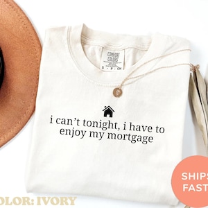 May include: Ivory-colored t-shirt with the text "i can't tonight, i have to enjoy my mortgage" in black. A gold necklace with a pendant is on the shirt. The shirt is labeled "Comfort Colors".