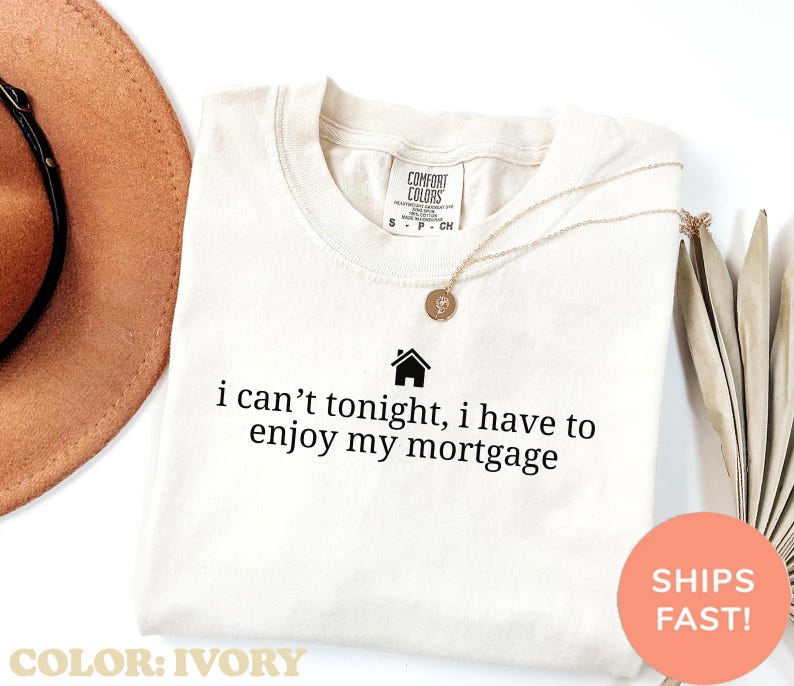 May include: Ivory-colored t-shirt with the text "i can't tonight, i have to enjoy my mortgage" in black. A gold necklace with a pendant is on the shirt. The shirt is labeled "Comfort Colors".