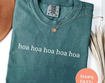 May include: Blue spruce Comfort Colors t-shirt with the text "hoa hoa hoa hoa hoa" in white. A gold-colored necklace with a pendant is draped on the shirt. The shirt is folded and ready to wear.
