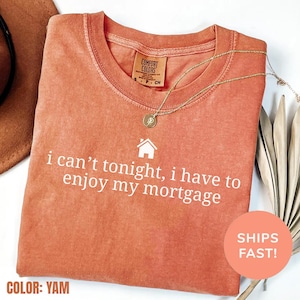 May include: A yam-colored Comfort Colors t-shirt with the text "i can't tonight, i have to enjoy my mortgage" in white. A gold necklace with a pendant rests on the shirt. The shirt is folded and has a small house graphic.