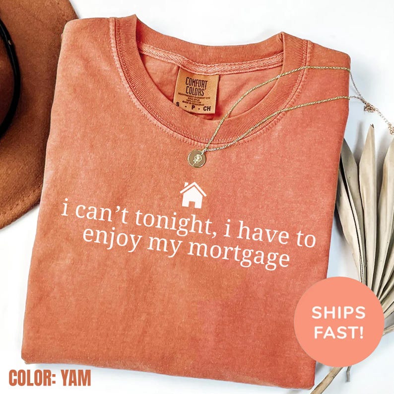 May include: A yam-colored Comfort Colors t-shirt with the text "i can't tonight, i have to enjoy my mortgage" in white. A gold necklace with a pendant rests on the shirt. The shirt is folded and has a small house graphic.