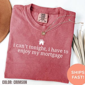 May include: Crimson-colored t-shirt with the text "i can't tonight, i have to enjoy my mortgage" in white. The shirt has a crew neck and a small tag at the top. A gold necklace with a pendant is draped across the shirt.