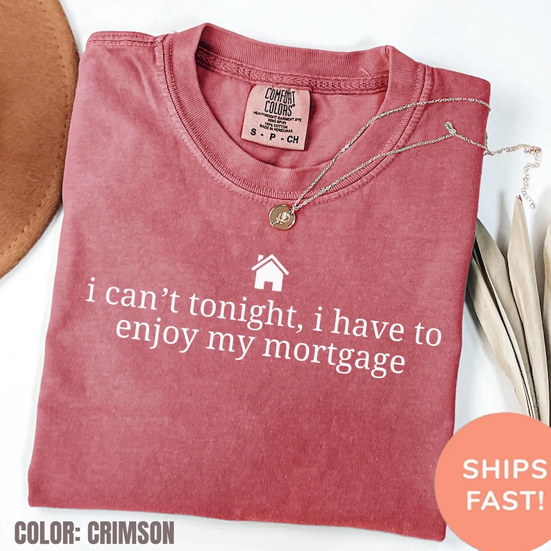 May include: Crimson-colored t-shirt with the text "i can't tonight, i have to enjoy my mortgage" in white. The shirt has a crew neck and a small tag at the top. A gold necklace with a pendant is draped across the shirt.
