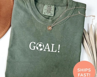 May include: A moss green t-shirt with the word "GOAL!" and a soccer ball graphic printed in white. The shirt has a crew neck and short sleeves. A gold necklace with a pendant is draped on the shirt. The shirt is labeled "Comfort Colors".