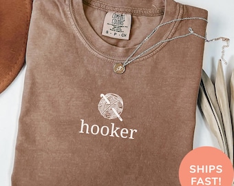 May include: Espresso-colored t-shirt with a white graphic of a yarn ball and crochet hook, with the word "hooker" below. A silver necklace with a small pendant rests on the shirt. The shirt is folded and has a Comfort Colors tag.