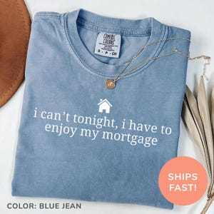 May include: A blue jean Comfort Colors t-shirt with the text "i can't tonight, i have to enjoy my mortgage" in white. A gold necklace with a pendant rests on the shirt. The shirt is folded and ready to wear.
