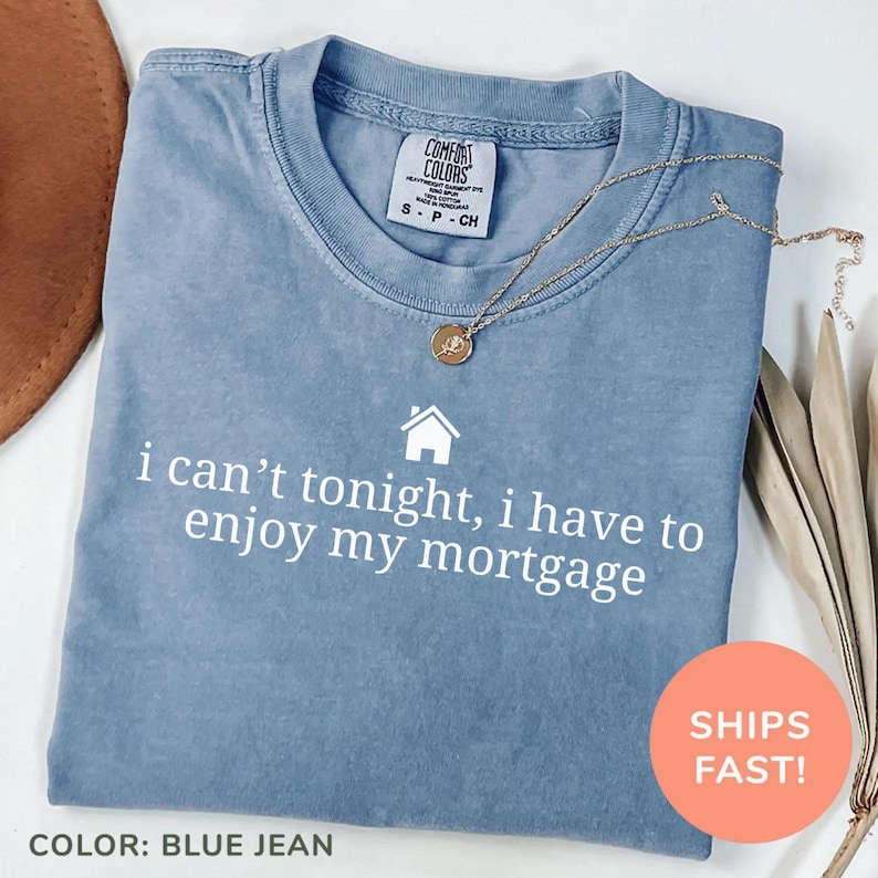 May include: A blue jean Comfort Colors t-shirt with the text "i can't tonight, i have to enjoy my mortgage" in white. A gold necklace with a pendant rests on the shirt. The shirt is folded and ready to wear.
