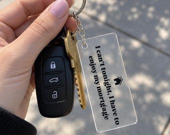 May include: A rectangular clear acrylic keychain featuring the black printed text I can't tonight, I have to enjoy my mortgage and a small house icon. The keychain is attached to a silver metal split ring along with a brass key and a black car key fob.