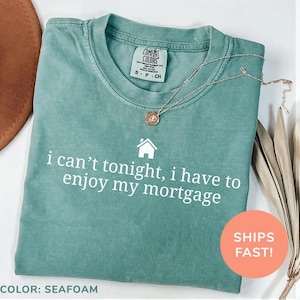 May include: Seafoam green Comfort Colors t-shirt with the text "i can't tonight, i have to enjoy my mortgage" in white. A gold necklace with a pendant rests on the shirt. The shirt is size small.