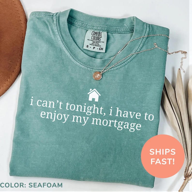 May include: Seafoam green Comfort Colors t-shirt with the text "i can't tonight, i have to enjoy my mortgage" in white. A gold necklace with a pendant rests on the shirt. The shirt is size small.