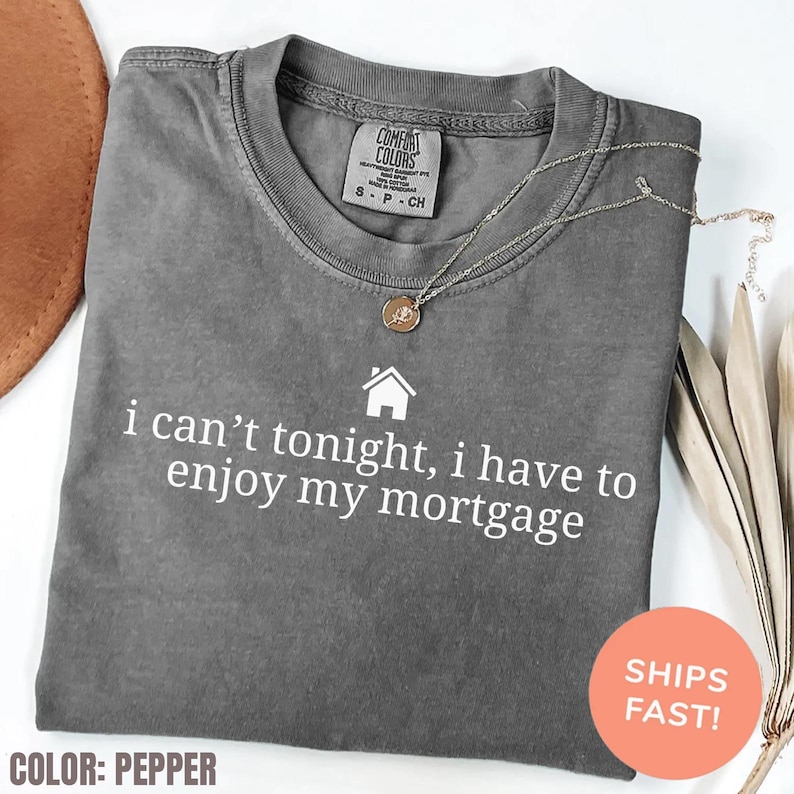 May include: Pepper-colored Comfort Colors t-shirt with the text "i can't tonight, i have to enjoy my mortgage" in white. A silver necklace with a pendant is draped on the shirt. The shirt has a crew neck.