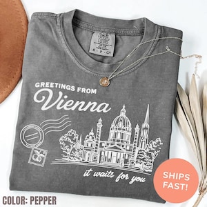 May include: A grey t-shirt with the words "GREETINGS FROM Vienna" and a city skyline illustration. The phrase "it waits for you" is also printed on the shirt. A gold necklace and a brown hat are also visible.