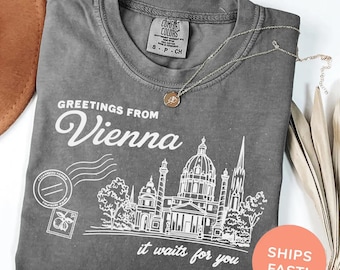 May include: A gray t-shirt with the text "GREETINGS FROM Vienna" and an illustration of a city skyline. The shirt also features the phrase "it waits for you". A gold necklace and a brown hat are also visible.