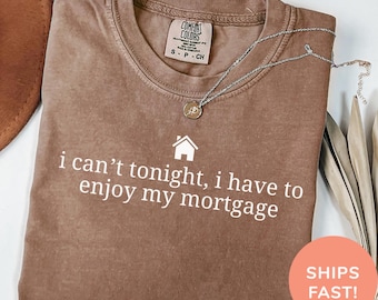 May include: Espresso-colored Comfort Colors t-shirt with the text "i can't tonight, i have to enjoy my mortgage" in white. The shirt has a crew neck and short sleeves. A silver necklace with a pendant is draped across the shirt.