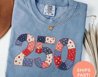 May include: A blue jean Comfort Colors t-shirt with the number "250" appliqued in a patchwork design of red, white, and blue fabrics. A gold necklace with a pendant rests on the shirt. The shirt is folded and displayed on a white surface.