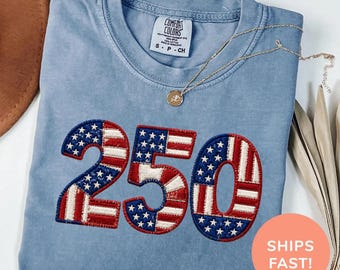 May include: A blue jean Comfort Colors t-shirt with the number 250 embroidered in red, white, and blue, resembling the American flag. A gold necklace with a pendant rests on the shirt. The shirt is folded and ready to wear.