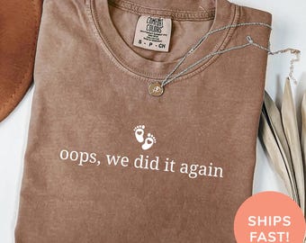 May include: Espresso-colored t-shirt with the text "oops, we did it again" and baby footprints. A silver necklace with a pendant rests on the shirt. The shirt is folded and ready to wear.