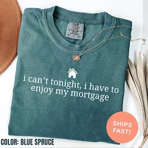 May include: Blue spruce Comfort Colors t-shirt with the text "i can't tonight, i have to enjoy my mortgage" in white. A gold necklace with a pendant rests on the shirt. The shirt is folded.