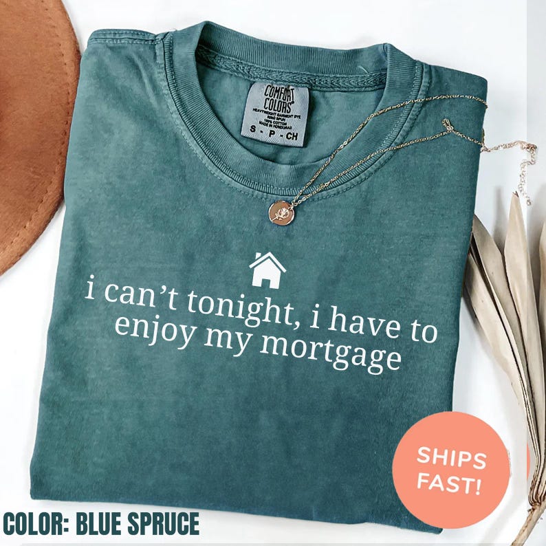 May include: Blue spruce Comfort Colors t-shirt with the text "i can't tonight, i have to enjoy my mortgage" in white. A gold necklace with a pendant rests on the shirt. The shirt is folded.