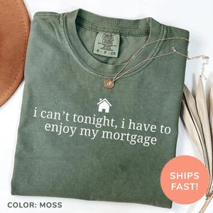 May include: A moss green Comfort Colors t-shirt with the text "i can't tonight, i have to enjoy my mortgage" in white. A gold necklace with a pendant rests on the shirt. The shirt is folded neatly.