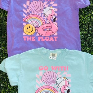 May include: Two short-sleeved t-shirts, one lavender and one light blue, with a graphic design. The design includes a pink flamingo float, rainbow, smiley face, and the text "GO WITH THE FLOAT".