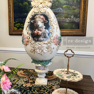 May include: A large, ornate egg-shaped decorative piece with a lamb portrait, floral accents, and a bow. The egg is cream-colored with gold and pink details. A two-tiered serving tray with floral designs is also present.