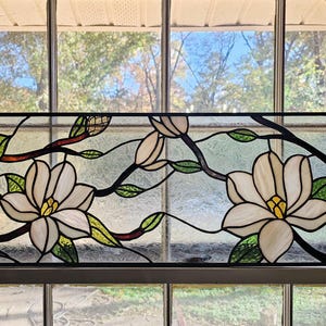 Pode incluir: Painel de janela de vitral com flores de magnólia brancas com centros amarelos, folhas verdes e galhos marrons escuros. O painel retangular está emoldurado em uma janela, com uma vista de árvores e céu visíveis através do vidro.