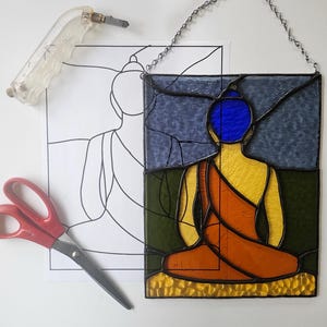 May include: A stained glass panel depicting a seated Buddha figure, featuring a blue head, yellow body, and orange robes. The panel is framed in black and hangs from a silver chain. A glass cutter, scissors, and a line drawing of the Buddha are also present.