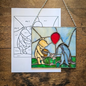 Op de afbeelding: Een gebrandschilderd raam met Winnie de Poeh en Biggetje. Winnie de Poeh houdt een honingpot vast en Biggetje houdt een rode ballon vast. Het gebrandschilderde raam is omlijst met een zilveren ketting.