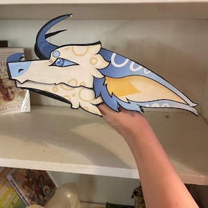 Puede incluir: Recorte de cabeza de dragón estilizado con una combinación de colores azul claro, amarillo y blanco. El dragón tiene cuernos grandes y curvos, un hocico largo y marcas detalladas en los ojos. El recorte se sostiene frente a una estantería con libros y una figura decorativa de caballo.