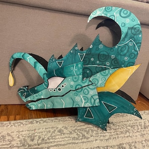 May include: A teal and turquoise dragon head sculpture with yellow accents. The artwork features a stylized design with geometric shapes and circular patterns. The dragon's eye is white, and a teardrop shape hangs from its snout.