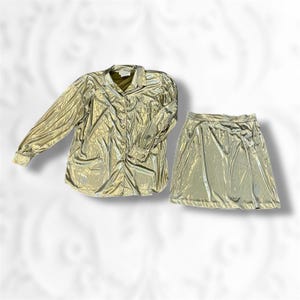 May include: A gold-colored, two-piece outfit consisting of a long-sleeved button-down shirt and a mini skirt. The fabric has a crinkled texture, giving it a metallic appearance. The shirt has a collar and buttoned cuffs. The skirt is a simple, A-line design.