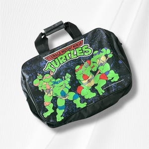 Peut inclure: Sac à bandoulière noir avec les Tortues Ninja. Le sac a une poignée et un graphisme des quatre tortues en poses d'action. Le fond est bleu foncé avec un motif éclaboussé.