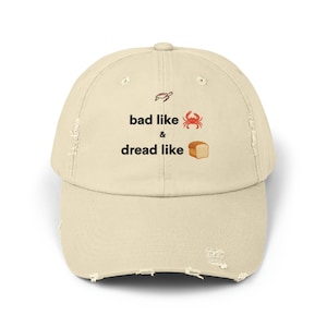 Puede incluir: Una gorra de béisbol desgastada de color beige con el texto "bad like & dread like" y emojis de una tortuga, un cangrejo y una hogaza de pan.