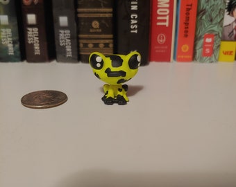 Puede incluir: Una pequeña figura de rana amarilla con manchas negras y ojos grandes y redondos. El juguete está sobre una estantería blanca, con una moneda a la izquierda y una fila de libros al fondo.