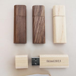 May include: Three wooden USB flash drives, two are dark brown and one is light brown. The light brown drive is open and shows a silver USB connector and the text "ikimi2023" engraved on the wooden surface.