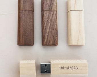Puede incluir: Tres unidades flash USB de madera, dos son de color marrón oscuro y una es de color marrón claro. La unidad flash de color marrón claro está abierta y muestra un conector USB plateado y el texto "ikimi2023" grabado en la superficie de madera.