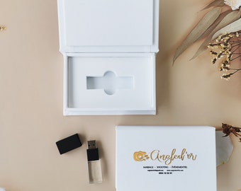 Könnte beinhalten: Eine weiße Box mit einem USB-Stick darin. Die Box ist geöffnet und der USB-Stick ist sichtbar. Der USB-Stick ist transparent und hat ein goldenes Etikett mit dem Text "Oz Angled'or" in goldenen Buchstaben. Der Text unter dem Logo lautet "Mariage - Shooting - Evenementiel" und "ozangledor.com" in kleinerer Schrift. Die Telefonnummer "0664 16 33 87" ist unter der Website-Adresse gedruckt.