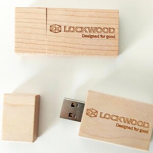 Puede incluir: Dos memorias USB rectangulares de madera clara con tapas extraíbles. Cada unidad presenta un logotipo grabado con láser con un icono geométrico, el texto LOCKWOOD y la frase Designed for good. Una unidad se muestra con la tapa retirada.