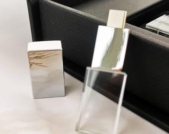 Könnte beinhalten: Zwei rechteckige, silberfarbene Parfümflakons mit weißen Deckeln. Einer steht aufrecht, der andere liegt. Die Flakons stehen auf einer weißen Oberfläche, mit einer schwarzen Box im Hintergrund.