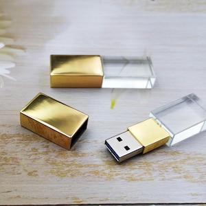Può includere: Chiavette USB dorate con corpi in cristallo rettangolari e trasparenti. Una chiavetta è aperta, rivelando il connettore USB. Le chiavette sono su una superficie di legno chiaro, con un fiore bianco sullo sfondo.